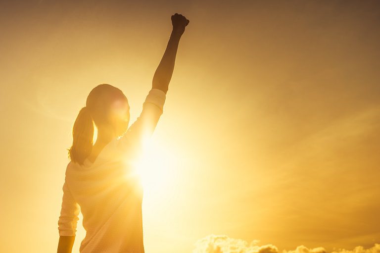 Person raising fist triumphantly against bright sunset light.